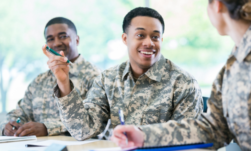 Two young men in military uniform smiling and talking during a classroom session, one of them holding a pen