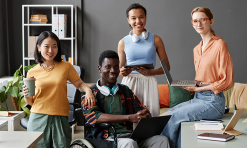 Group of diverse young adults in a casual workspace, smiling at the camera. One individual is seated in a wheelchair, and others are holding laptops and tablets, representing inclusion and collaboration.