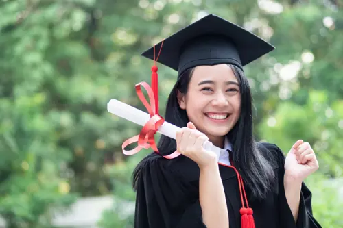 Smiling girl in graduation robes holding a diploma tied with a red ribbon, looking proudly into the camera