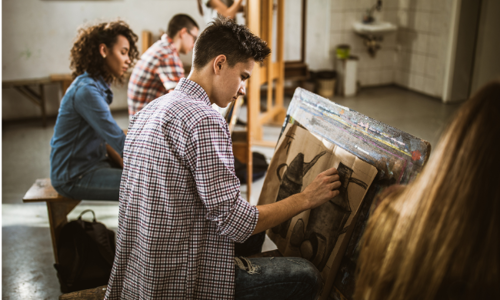 A high school student sitting at an easel sketching a still-life drawing in an art class, with other students working in the background