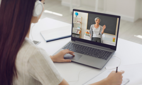 A student attending an online class at home, watching a teacher on a laptop screen and taking notes with headphones on