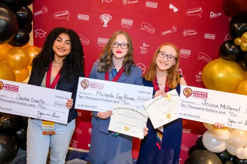 Three young female students posing in front of a photo wall with NSHSS branding, holding their scholarship checks and NSHSS member certificates