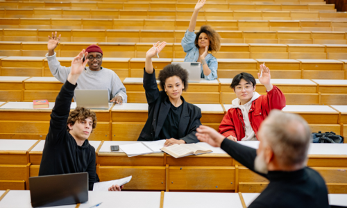 College students in a lecture hall raising their hands to participate, with laptops and books open in front of them, showing an engaged and diverse classroom setting