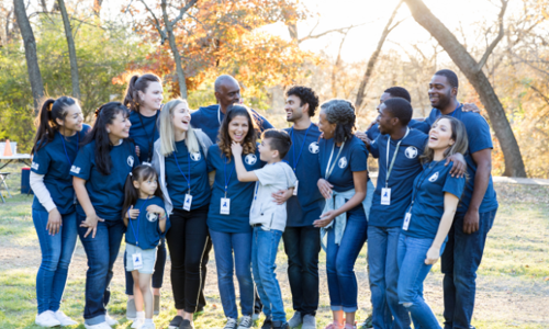 A diverse group of volunteers, including adults and children, wearing matching blue shirts and smiling together outdoors in a park