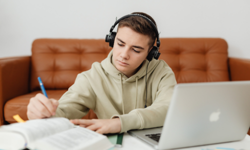 A young student wearing headphones, writing in his notebook while sitting at a desk with a laptop on it