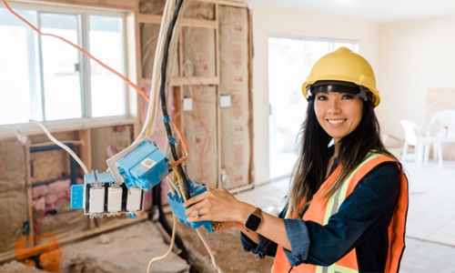 A woman in a construction site wearing a hard hat and safety vest, holding electrical wires and smiling at the camera