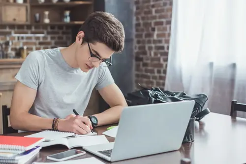 A young man is writing in a notebook sitting by the desk