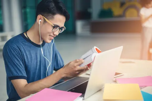 A young man studying, holding a workbook and listening to the audio in his headphones
