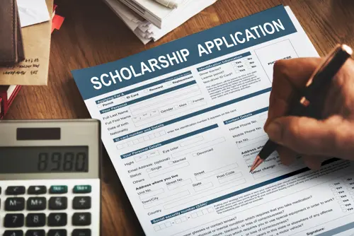 A hand filling out a form labeled "Scholarship Application"